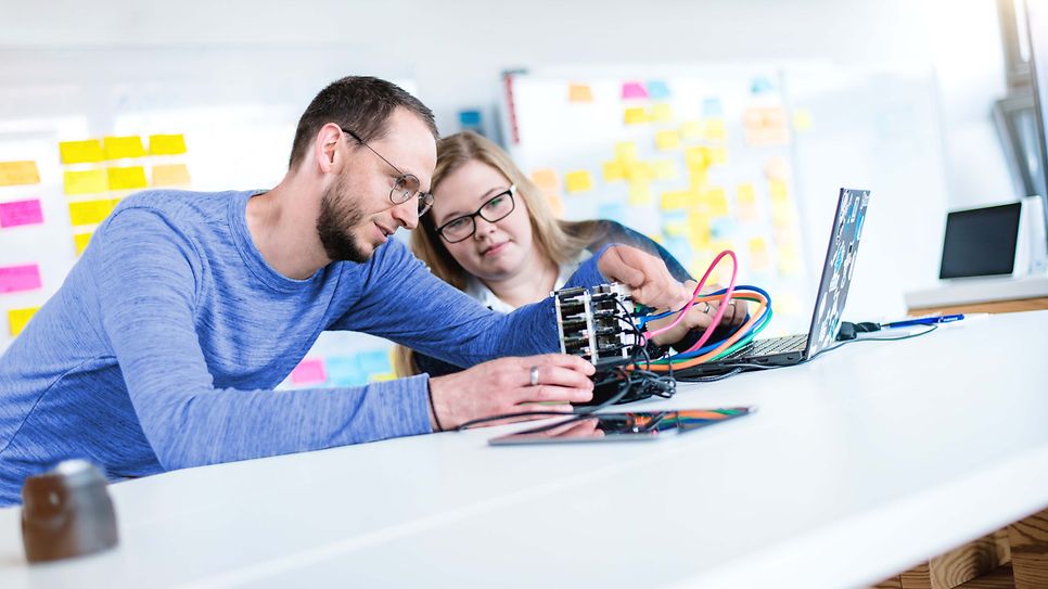 Ausbilder erklärt Auszubildende ein elektronisches Gerät was auf dem Tisch liegt
