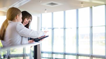 Zwei Auszubildende stehen am Geländer und sehen sich etwas auf einen Tablet an.