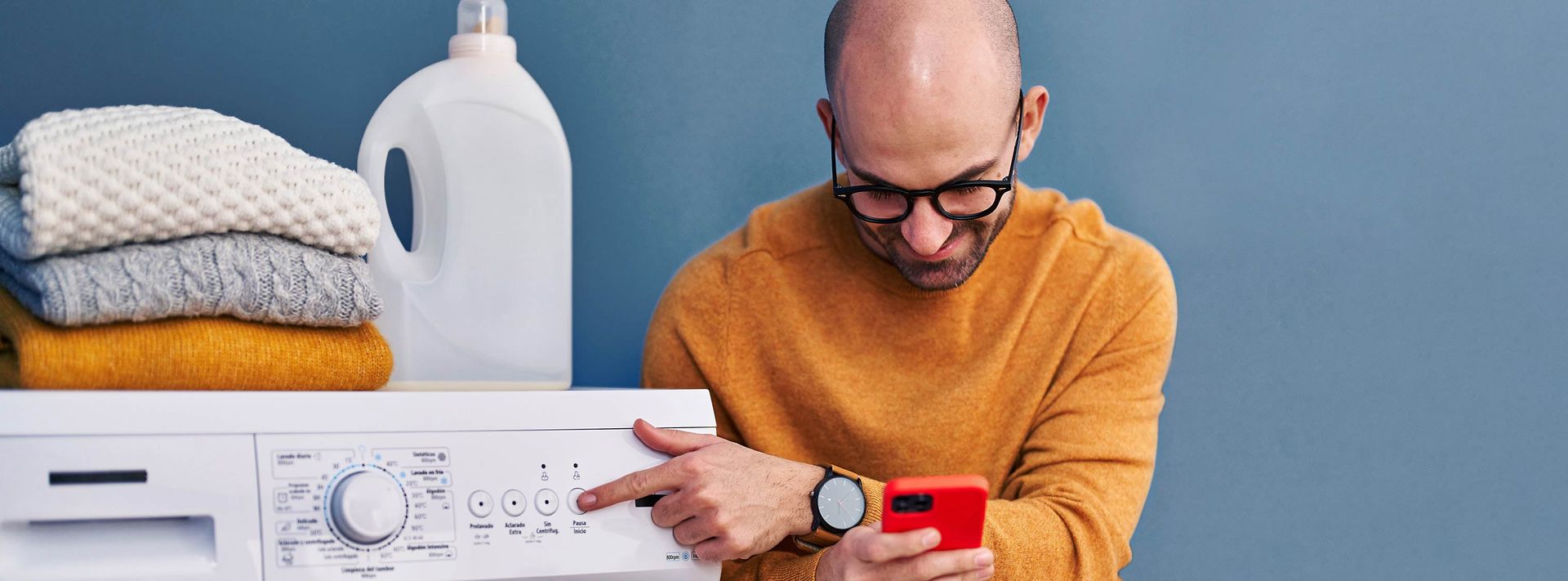 Mann mit Brille schaut auf sein Handy während er seine Waschmaschine bedient.